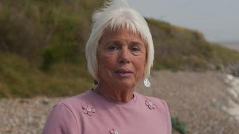 Maureen Jones, a woman who is standing on the beach at Thorpeness in Suffolk. She is wearing a pink T-shirt with pink flowers on it. She is looking directly at the camera and smiling and has white short hair. 