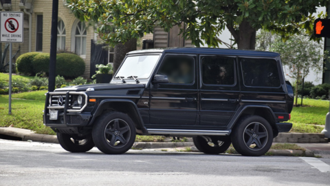 A a black Mercedes Benz G Wagon on a road ear a tree, grass verge and buildings in the background