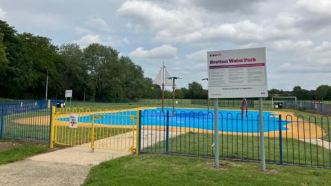 A small paddling pool surrounded by blue paving and a fence. There is a building next to it with pool rules written on the side.