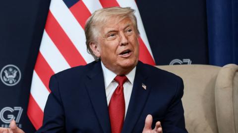 Donald Trump wearing a red tie and speaking in front of the US flag
