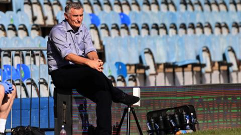 Tony Mowbray, wearing a lilac shirt and black trousers, sits on the advertising boards at London Road Stadium while managing Blackburn Rovers against Peterborough United in 2022