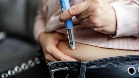 A woman, whose face and upper torso is not shown, injects a weight-loss drug via an injection pen into her abdomen, with the belt of her blue jeans unbuckled and her pink jumper lifted up slightly