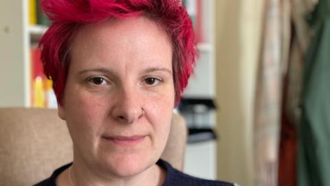 Sarah Croney sits in her office on a beige coloured chair. She has dark brown eye and pink hair. She is wearing a navy blue jumper.