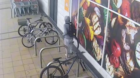 A CCTV image showing a man walking alongside a row of parked bikes while holding a wheel.