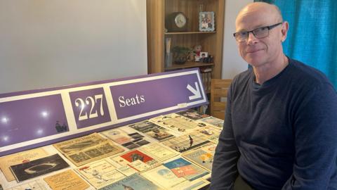 Graham Cooksley sat in his kitchen in front of a selection of his collection which is displayed out on the table. They are a lots of programmes, tickets and books as well as a big purple original Wembley stadium seat sign.
