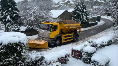 Yellow snow plough/gritting lorry makes its way along a snowy street leaving clear road behind it