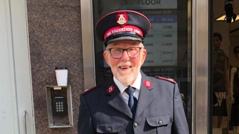 Major John Mott wearing a black Salvation Army uniform and cap that reads, "The Salvation Army". He is standing outside a shop. He has black glasses and a white beard and is smiling.