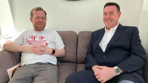 Two men sit on a sofa, smiling at the camera. One is wearing a T-shirt and shorts, the other a dark suit and white shirt.