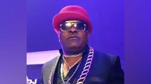 A grab from a video of Longue Longue at a concert. He's wearing gold chains, dark glasses and a red bowler hat