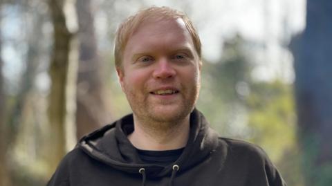 David Brody has short red/blond hair and stubble. He is smiling and wearing a black hoodie. There are trees in the background.