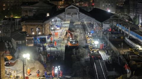 A railway station at night with several people dressed in orange on the platforms along with machinery. The platform is lit up by floodlights. 