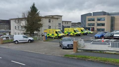 A series of buildings make up Letterkenny Hospital. Three ambulances are parked in a car park. Several cars are also parked. It's a dull cloudy day. 