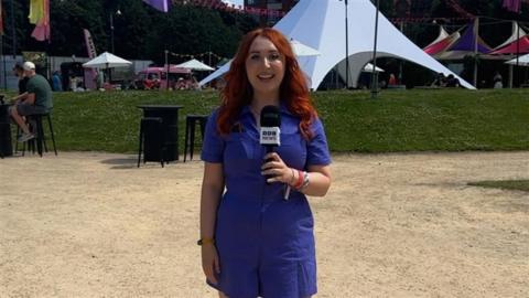 A woman in a blue dress with red hair standing in a sunny festival arena