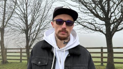 A man stood in a park, on a frosty foggy day. He is wearing a black denim jacket, a white hoody, a black cap and black sunglasses. He has tattoos on his neck. He is stood in front of three bare trees