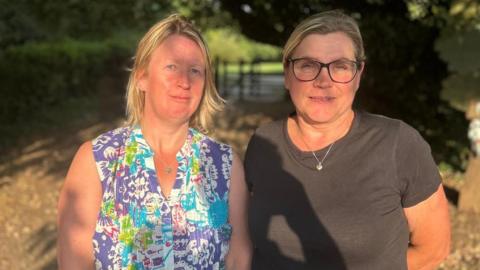 Two blonde women stand looking at the camera. The woman on the left is wearing a blue patterned top. The woman on the right has glasses and a dark coloured tshirt with necklace.