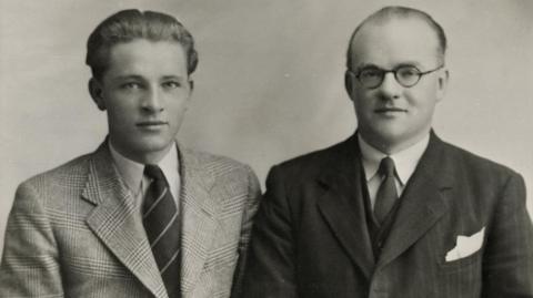 Two men wearing suits in a black and white photograph. The older man is wearing a darker suit with glasses and has a receding hairline.