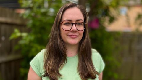 Erin wearing a green top and glasses. She is looking direct at the camera and standing in a garden. She has long brown hair hanging to chest level.