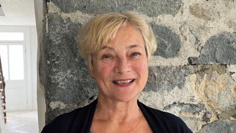 A woman with short blonde hair, smiling at the camera, standing against a grey brick wall beside a hallway