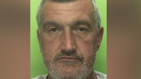 A white man with stubble stares at the camera for a police mugshot. He has greying hair and thin lips.