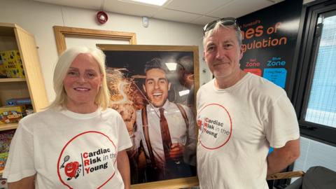 Lesley and Robin Perkins, both wearing a white T-shirt with the logo Cardiac Risk in the Young, stand inside a converted classroom in front of a large picture of their son, Dan who is holding a sparkler.