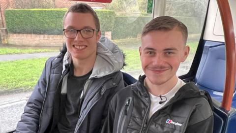 This picture shows best friends Tyler and Alfie on board a bus to York city centre. They are sitting next to each other on the lower deck, towards the back of the bus. Tyler is wearing glasses and a grey coat. Alfie is wearing a black hoodie and a white T-shirt. They are both smiling.