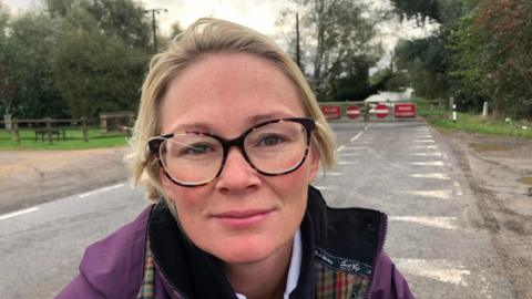 Stacey Buchanan with short, blonde, swept-back hair. She has large horn-framed glasses and is wearing a white shirt under a purple tartan-lined overcoat. Behind her is the Earith causeway, with red 'road closed' and 'no entry' signs. The flood water is lapping the road and a fallen tree can be seen a few metres into the flood.