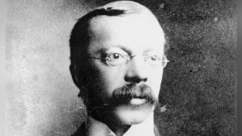 A black and white portrait-style photograph of a man with a moustache which curls at the ends, a side parting, and thin framed glasses. His face is angled to the left and he is gazing beyond the camera.