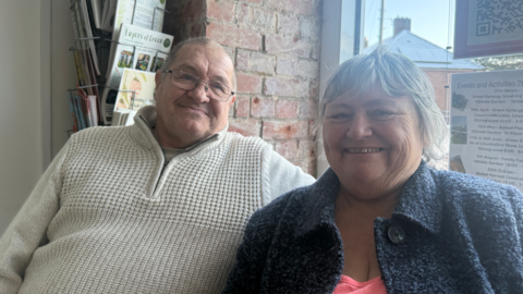 Alan Thompson is wearing a white jumper and has glasses with black edged rims. He is sat next to Jane Innes who is wearing a blue jacket and is smiling. Behind them is a red bricked wall and a window.