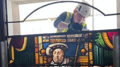 A large and colourful stained glass panel which shows Henry VIII handing a charter to Sir Thomas Vicary. The panel is full of rich yellows, blues and reds. Behind the panel is a man wearing a hard hat and high vis jacket, appearing to fix the top of the panel. It is unclear what tool he is using.