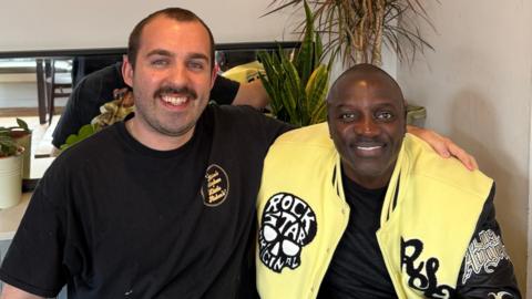 Aidan Jackson wearing a black t-shirt poses with Akon who is wearing a yellow, black and gold jacket. The jacket has a logo which reads "Rock Star Original". 