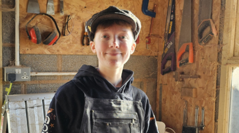 Elizabeth, standing in her workshop smiling to camera, wearing a black apron, and dark blue hoody, both dirty from her work. She is also wearing a dark green flat cap and there are carpenter's tools hanging above the bench behind her.