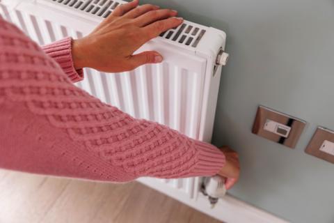 A woman wearing a pink woolly jumper is touching a radiator with her left hand while adjusting the knob on the radiator with her right hand, 