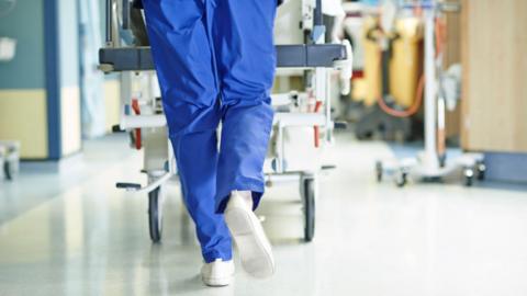 A hospital worker in blue trousers with white shoes is walking in a corridor, pushing a hospital trolley in front of them