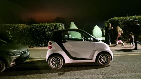 A black and white Smart car is parked across a dropped kerb outside a bowls club. There is another car behind it and pedestrians walking past on the pavement. There are large bushes either side of the entrance, which is pictured at night.