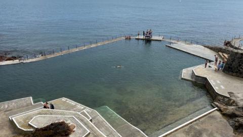 A swimming pool is built into the coastline, with one wall adjacent to the sea. There is a graduated sloped down to the pool and the words ' no diving from edge' can just be seen. There are a few people in the water and a handful are standing besides the pool, including a child with a water wing.