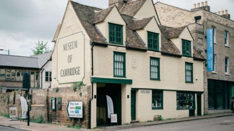 A large end of terrace building is in the middle. It is painted a light yellow and has green framed windows. There is a sign on the end of the building, which is on the left, that says MUSEUM OF CAMBRIDGE. 