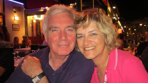 The image shows two people sitting closely together at an outdoor restaurant or café during the evening. They are seated at a table with white tablecloths, and there are glasses and plates visible. One person is wearing a dark short-sleeved shirt and a wristwatch, while the other is wearing a bright pink collared shirt with a small embroidered logo. The background features other diners, warm ambient lighting, and a street scene with illuminated buildings and lights.