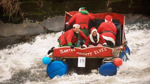 A homemade raft sailing on a river with four people on it. The raft is made from blue drums, wooden panels, a black sofa and red painted wood carved in the shape of a sleigh. The words 'Santas's naughty elves' is painted on the back of the raft. The four people are knelt on the raft dressed in Santa outfits. The waves are crashing and there are large stones and bushes on the bank.
