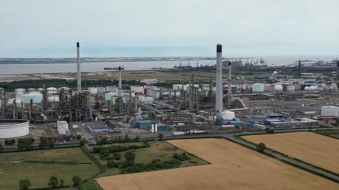 A drone view of the Prax Lindsey Oil Refinery. A cluster of metal structures rising to the grey sky. Green and yellow stubble fields can be seen. 