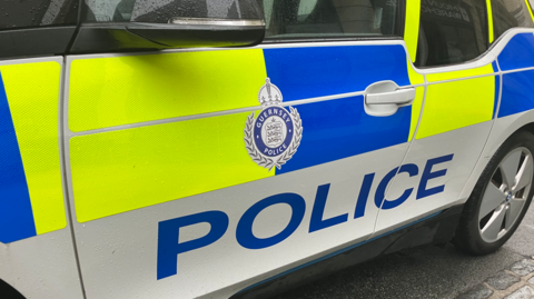 The side of a police car. There is the Guernsey Police logo, blue letters which read 'Police' and t