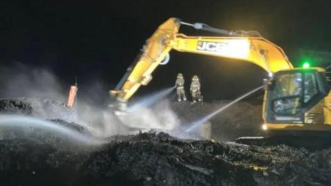 A yellow crane can be seen to the right, its arm out over a sea of scorched and blackened waste, while the beams of its light in the dark show two firefighters in yellow high-viz using a hose to help dampen the fire. Smoke is seen floating upwards from the scene. 