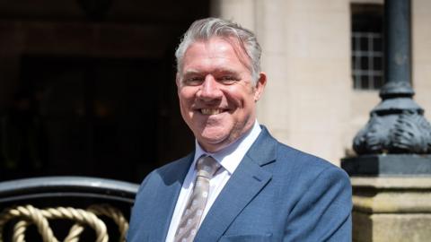 Anthony Screen is standing by a light-coloured stone building with railings in front of it. He is wearing a grey suit with a white shirt and a brown tie and is smiling at the camera.