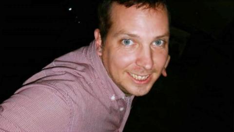 A man is leant forward into the middle of the frame, and is looking at the camera. He is smiling and wearing a red shirt. The background is black.