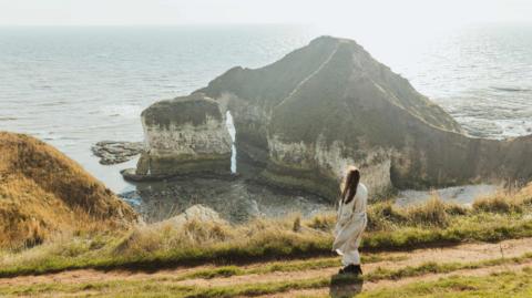 A woman with long dark hair walking along a grassy coastal path with sea stacks visible in the background.