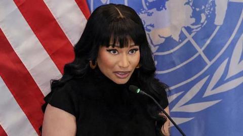 Nicki Minaj with long black hair and bangs, standing at a podium. She is dressed in a fitted black outfit. Behind her, two large flags hang side by side, one is the US  flag and the other has the UN emblem on a blue background