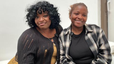 Two women are sitting next to each other and smiling. The woman on the left is wearing a black cardigan, a yellow top and has black curly hair. The woman on the right is wearing a plaid jacket and a black top.