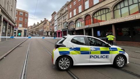 A cordon in Cheapside, Nottingham city centre. Police tape blocks tram lines and a police car is parked across the road