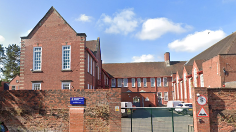 A red-brick school building behind a red-brick wall and green fence, with a blue sign on the wall.