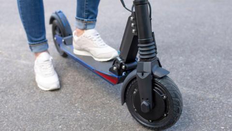 A person with a foot on an e-scooter, that is black, they are wearing white trainers and blue jeans 