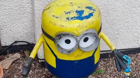 The gas canister is decorated as a minion and is looking up at the camera. He is stood in front of a house. 